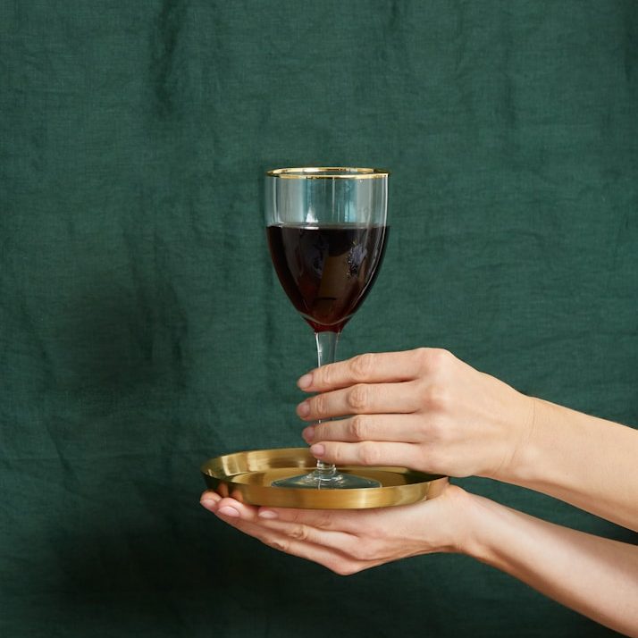 Händehaltung eines Weinglases auf einem goldenen Tablett vor grünem Hintergrund.